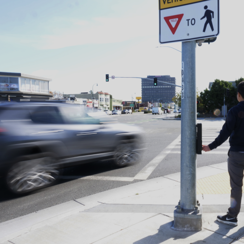 A pedestrian waits for traffic along a busy stretch of El Camino Real in San Mateo
